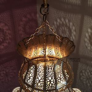 May include: A gold Moroccan pendant light with intricate cutouts and a scalloped top. The light is hanging from a chain and casts intricate shadows on the wall.