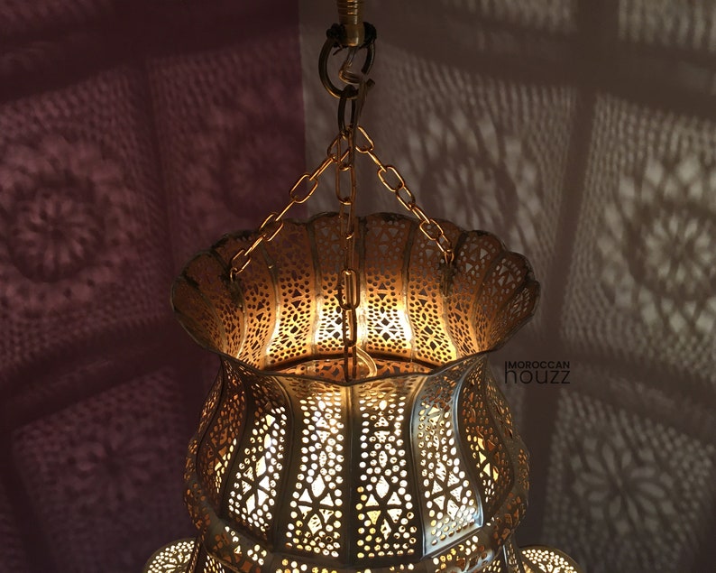 May include: A gold Moroccan pendant light with intricate cutouts and a scalloped top. The light is hanging from a chain and casts intricate shadows on the wall.