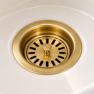 Puede incluir: Un desagüe de fregadero circular de latón cepillado con un pomo central y una serie de ranuras verticales espaciadas uniformemente para el drenaje del agua, encajado en una superficie de lavabo de cerámica blanca lisa.