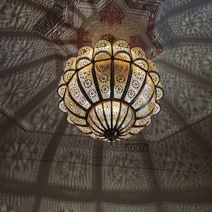 May include: A large, round, gold-colored Moroccan pendant light fixture with intricate cut-out designs. The light fixture is hanging from a ceiling with a white background. The light fixture casts a shadow on the ceiling.