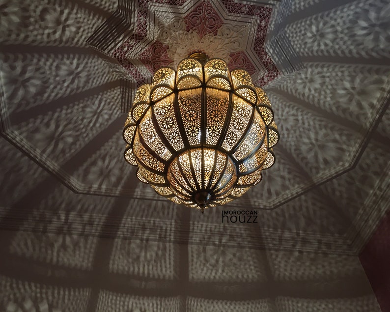 May include: A large, round, gold-colored Moroccan pendant light fixture with intricate cut-out designs. The light fixture is hanging from a ceiling with a white background. The light fixture casts a shadow on the ceiling.