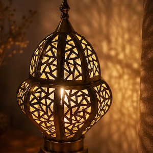 May include: A decorative brass lantern with a geometric cut-out design. The warm light from within casts intricate shadows on the surrounding surface. The lamp has a pointed top and a rounded base, creating a cozy ambiance.