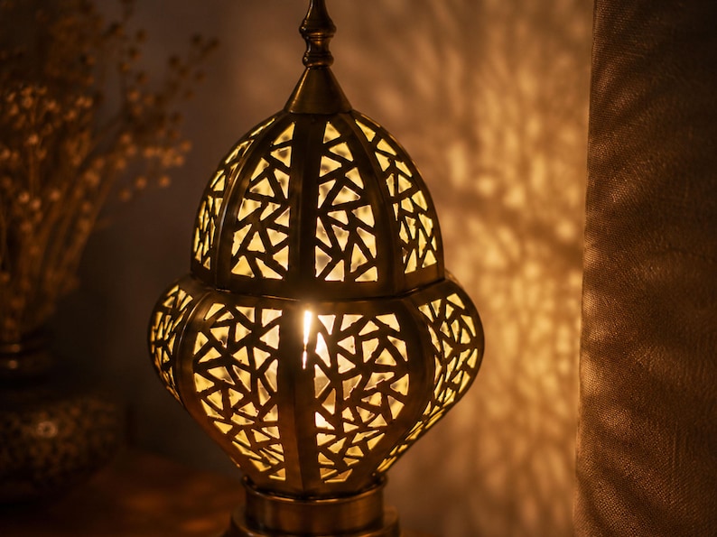 May include: A decorative brass lantern with a geometric cut-out design. The warm light from within casts intricate shadows on the surrounding surface. The lamp has a pointed top and a rounded base, creating a cozy ambiance.