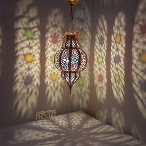 May include: A gold Moroccan pendant light with colorful glass accents casts intricate shadows on the wall. The light is hanging from a chain and has a round, openwork design.