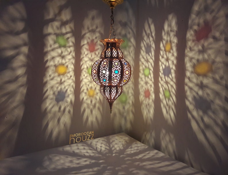 May include: A gold Moroccan pendant light with colorful glass accents casts intricate shadows on the wall. The light is hanging from a chain and has a round, openwork design.