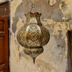 May include: A brass hanging lantern with intricate cut-out designs. The lantern is round with a pointed bottom and a decorative top. The lantern is hanging from a chain against a textured wall.