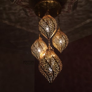 Może przedstawiać: Złota marokańska lampa wisząca z czterema ozdobnymi, liściastymi kloszami. Lampa jest zawieszona na suficie, a klosze są oświetlone.