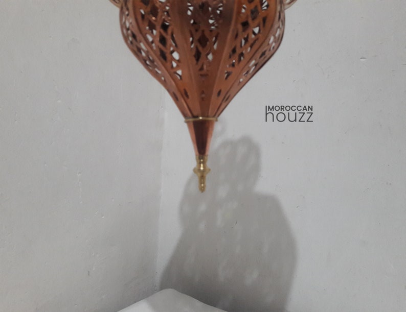 May include: A copper Moroccan lantern with intricate cut-out designs. The lantern is hanging from the ceiling and has a pointed tip at the bottom.