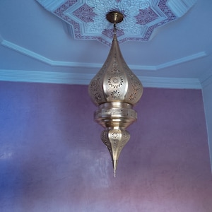 May include: A gold-coloured metal pendant light fixture with intricate cut-out designs hangs from a white ceiling with a decorative pattern. The light fixture is shaped like a teardrop and has a pointed bottom.