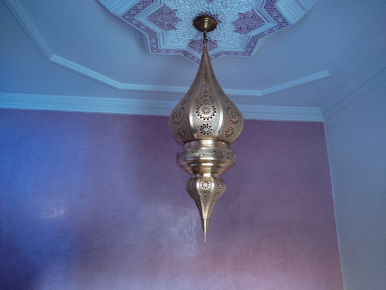 May include: A gold-coloured metal pendant light fixture with intricate cut-out designs hangs from a white ceiling with a decorative pattern. The light fixture is shaped like a teardrop and has a pointed bottom.