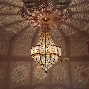May include: A large, ornate gold chandelier with intricate cutouts and a geometric design hangs from a ceiling with a pink and white patterned design. The chandelier casts shadows on the walls.