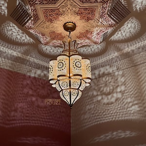 May include: A gold Moroccan lantern with intricate cutouts hangs from a ceiling with a pink and white patterned design. The lantern casts intricate shadows on the walls.