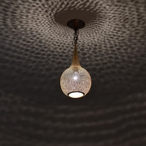 May include: A decorative pendant light fixture with a perforated metal shade. The light is suspended from the ceiling by a chain. The shade is a warm gold color, and the light casts a patterned shadow on the ceiling.
