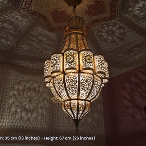 May include: A large, ornate, gold-colored metal chandelier with intricate cut-out designs. The chandelier is hanging from a ceiling and casts intricate shadows on the wall. Width: 35 cm (13 inches) - Height: 67 cm (26 inches)