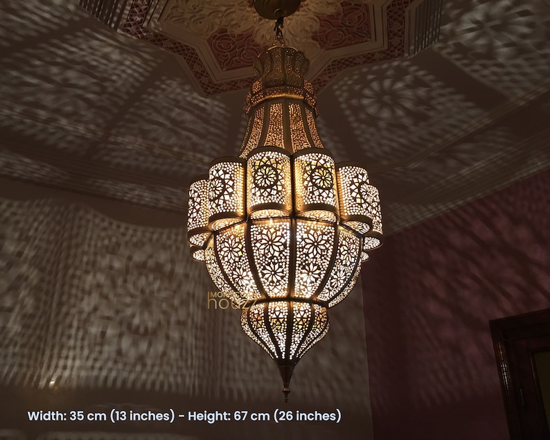 May include: A large, ornate, gold-colored metal chandelier with intricate cut-out designs. The chandelier is hanging from a ceiling and casts intricate shadows on the wall. Width: 35 cm (13 inches) - Height: 67 cm (26 inches)