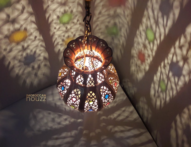 May include: A Moroccan-style hanging lantern with intricate metalwork and colorful glass accents. The lantern casts a warm glow and creates beautiful shadows on the ceiling.