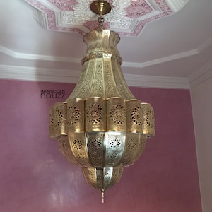 May include: A large, ornate gold-colored metal chandelier with intricate cut-out designs. The chandelier is hanging from a white ceiling with a pink and white decorative pattern.