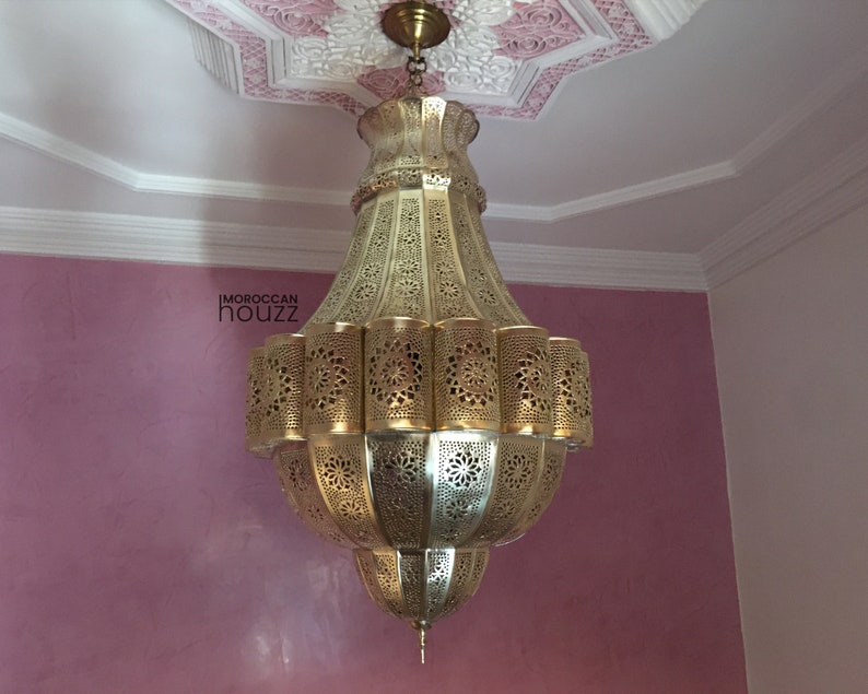 May include: A large, ornate gold-colored metal chandelier with intricate cut-out designs. The chandelier is hanging from a white ceiling with a pink and white decorative pattern.