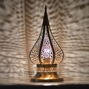 May include: A decorative brass lamp with a pierced, teardrop-shaped shade. The lamp has a detailed floral pattern and a pointed top. The base is round and the lamp casts intricate shadows on the background. The text "Moroccan Houzz" is visible on the base.