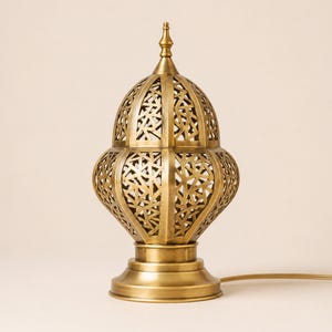 May include: A gold-colored, ornate table lamp with a domed, pierced shade. The lamp has a decorative, geometric pattern and sits on a round base. A power cord extends from the base.