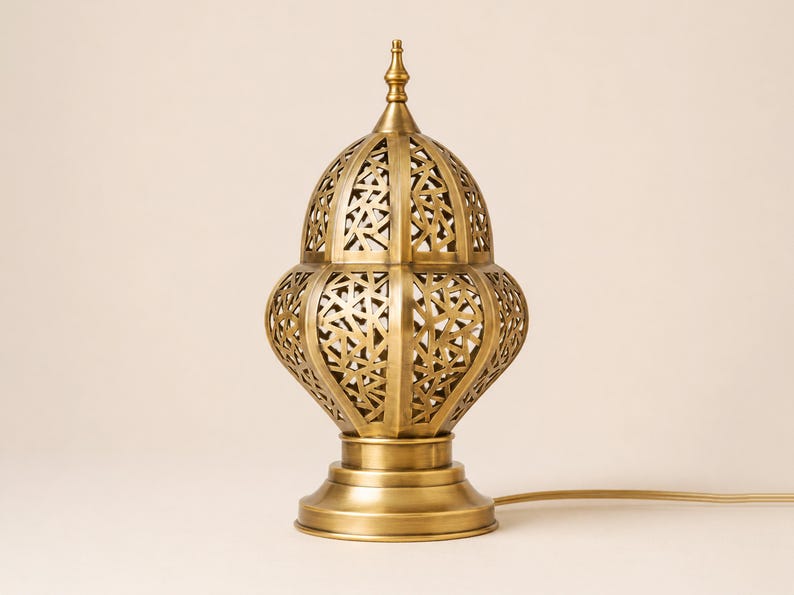 May include: A gold-colored, ornate table lamp with a domed, pierced shade. The lamp has a decorative, geometric pattern and sits on a round base. A power cord extends from the base.