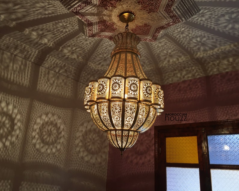 May include: A large, ornate, gold-colored chandelier with intricate cut-out designs hangs from a ceiling with a pink and white patterned design. The chandelier casts a shadow on the wall below.