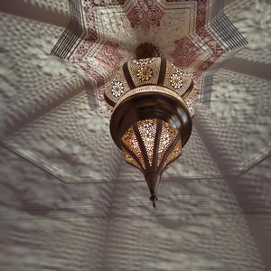 May include: A gold-toned metal ceiling light fixture with intricate cut-out designs. The light fixture is hanging from a white ceiling with a decorative pattern.