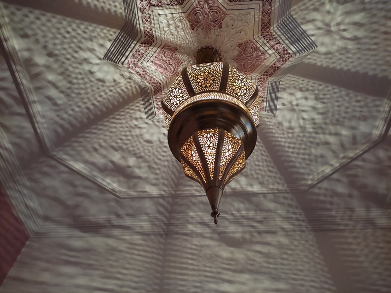 May include: A gold-toned metal ceiling light fixture with intricate cut-out designs. The light fixture is hanging from a white ceiling with a decorative pattern.