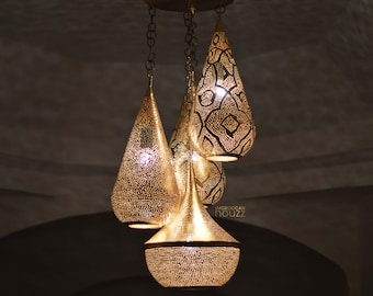 May include: Three gold-coloured, Moroccan-style pendant lights suspended from chains. Each light features a unique, intricate pattern of cut-out designs, creating a warm glow. The lights are set against a dark background.