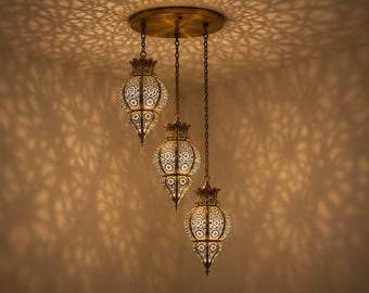 May include: A three-light pendant fitting featuring brass-toned metal lanterns with intricate, laser-cut floral patterns. Each lantern hangs from a central circular ceiling mount by a metal chain, casting detailed geometric shadows on the walls.