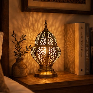 May include: A decorative brass lamp with a geometric pattern, casting intricate shadows. The lamp sits on a wooden surface next to a small vase with greenery and a stack of books. A small, round container is also visible.