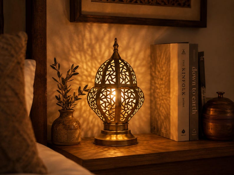 May include: A decorative brass lamp with a geometric pattern, casting intricate shadows. The lamp sits on a wooden surface next to a small vase with greenery and a stack of books. A small, round container is also visible.