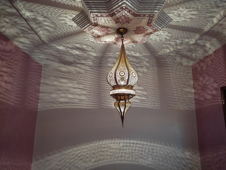 May include: A gold and white Moroccan pendant light fixture with intricate cutouts hangs from a ceiling with a star-shaped design. The light casts a pattern of shadows on the wall.