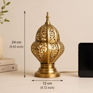 May include: A gold-colored metal table lamp with an intricate, openwork design. The lamp is 24 cm (9.45 inches) tall and 12 cm (4.72 inches) wide. A black smartphone is next to the lamp. The lamp is on a white surface.