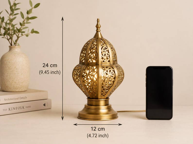 May include: A gold-colored metal table lamp with an intricate, openwork design. The lamp is 24 cm (9.45 inches) tall and 12 cm (4.72 inches) wide. A black smartphone is next to the lamp. The lamp is on a white surface.