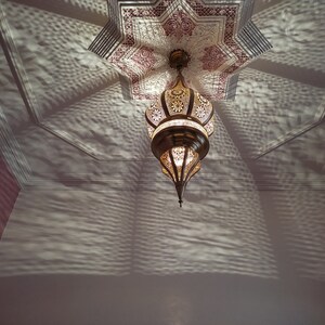 May include: A gold metal Moroccan-style chandelier with intricate cut-out designs hangs from a ceiling with a pink and white star-shaped pattern. The chandelier casts a shadow on the ceiling.