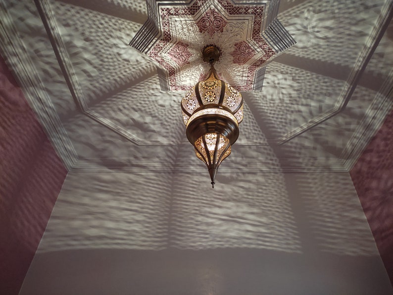 May include: A gold metal Moroccan-style chandelier with intricate cut-out designs hangs from a ceiling with a pink and white star-shaped pattern. The chandelier casts a shadow on the ceiling.
