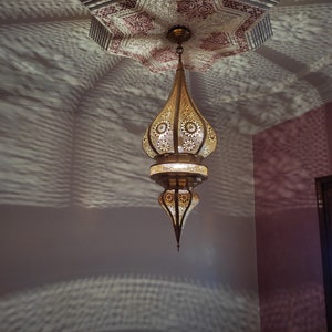 May include: A gold metal Moroccan pendant light fixture with intricate cut-out designs hangs from the ceiling. The light fixture casts a patterned shadow on the wall.