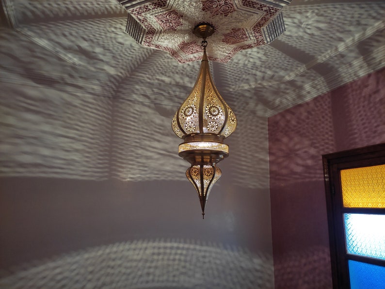 May include: A gold metal Moroccan pendant light fixture with intricate cut-out designs hangs from the ceiling. The light fixture casts a patterned shadow on the wall.