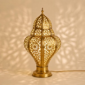 May include: A golden-colored, ornate table lamp with a detailed, geometric pattern. The lamp has a bulbous base, a tiered body, and a pointed top. The light shines through the cut-out design, casting decorative shadows.