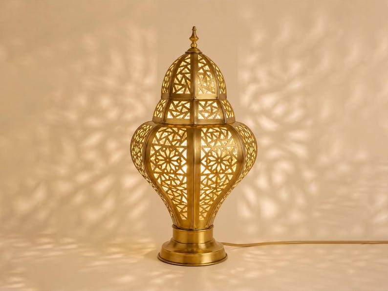 May include: A golden-colored, ornate table lamp with a detailed, geometric pattern. The lamp has a bulbous base, a tiered body, and a pointed top. The light shines through the cut-out design, casting decorative shadows.