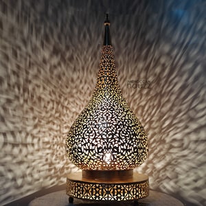 May include: A decorative, gold-colored metal lamp with a pierced design, casting intricate light patterns on the wall. The lamp has a teardrop shape with a pointed top and sits on a round base. The text "IMOROCCAN HAUZZ" is visible.