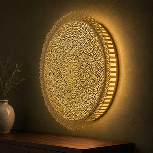 May include: A circular wall-mounted light fixture featuring an intricate, laser-cut gold metal filigree overlay with a dense, repeating floral pattern. The fixture emits a warm, soft glow from behind the metalwork, casting light onto the wall.