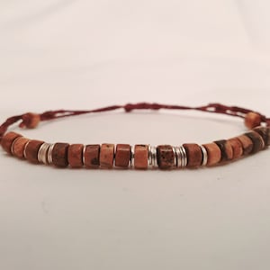 Op de afbeelding: Een armband met vierkante, roodbruine houten kralen en zilverkleurige metalen ringen. De armband is geregen op een donkerrood koord, met houten kralen aan elk uiteinde. Een eenvoudige, natuurlijke stijl.
