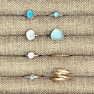 May include: A display of rings with various stones and materials, including a ring with a blue stone, a ring with a white shell, and a ring with a gold band.