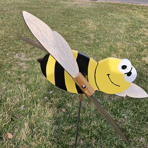 May include: A wooden bee wind spinner with yellow and black stripes, a smiling face, and large white eyes. The bee has two wings that are painted to look like wood grain.