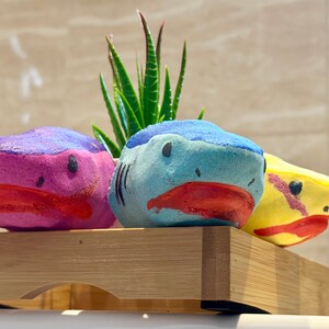 All three bath bombs are presented closer to see all the details. Blue shark in the middle, pink shark in the left side and the yellow shark in the right.