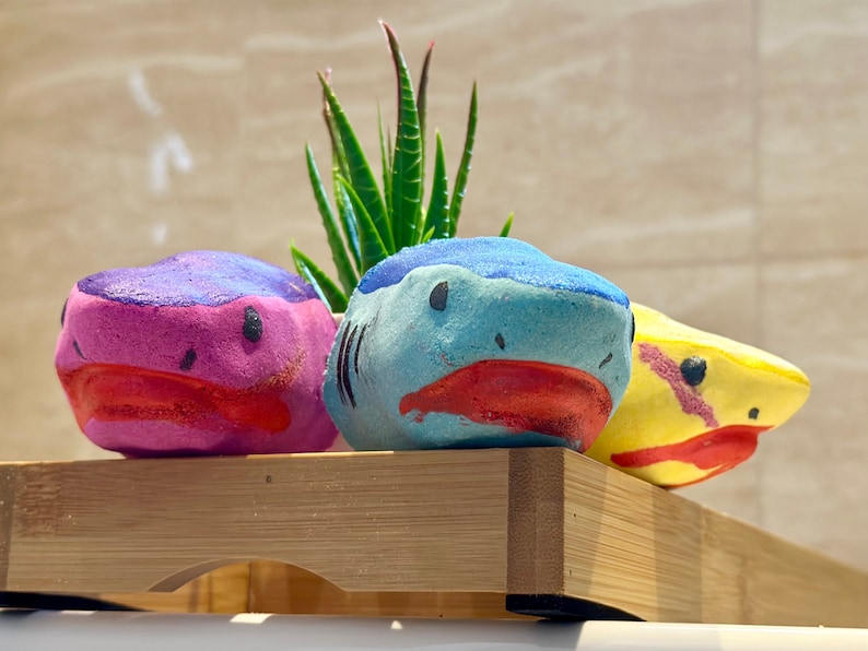 All three bath bombs are presented closer to see all the details. Blue shark in the middle, pink shark in the left side and the yellow shark in the right.