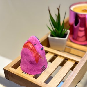 Handmade pink shark bath bomb resting over the bathtub. The bath bomb features a cute, hand-painted shark face with vibrant pink colour, perfect for a fun and fizzy bath time experience.