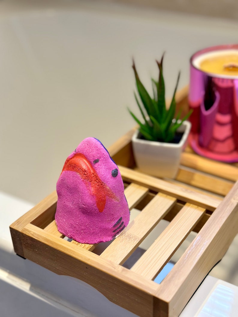 Handmade pink shark bath bomb resting over the bathtub. The bath bomb features a cute, hand-painted shark face with vibrant pink colour, perfect for a fun and fizzy bath time experience.